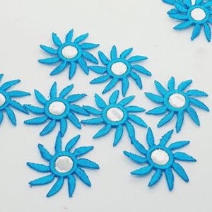 Puede incluir: Una colección de pequeños parches de flores azules bordadas con centros blancos. Los parches están dispuestos sobre una superficie blanca, mostrando su intrincado diseño de pétalos y detalles centrales reflectantes. Estos parches decorativos son adecuados para la decoración de telas.