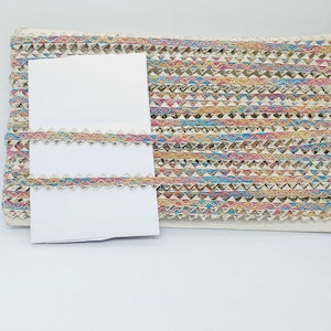 Puede incluir: Un rollo de ribete decorativo con un patrón geométrico repetitivo. El ribete presenta un borde plateado metálico con un diseño de ondas coloridas en tonos rosa, amarillo, azul y dorado. Se muestran dos pequeñas muestras del ribete en tarjetas blancas.