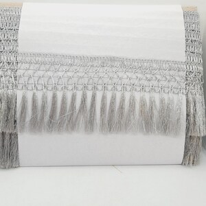 May include: Silver fringe trim with a delicate, woven pattern. The fringe is approximately 1 inch long and has a soft, silky texture.