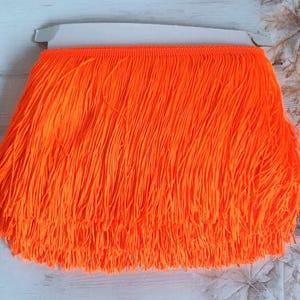 Puede incluir: Una cinta de flecos de color naranja brillante con una textura lisa. Los flecos tienen aproximadamente 2,5 cm de largo.