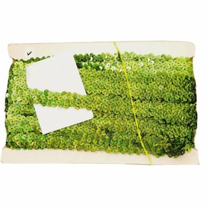May include: A roll of bright green sequins, tightly packed together, creating a textured surface. The sequins are a vibrant green colour. The roll is held together with a yellow string and is on a cardboard backing.