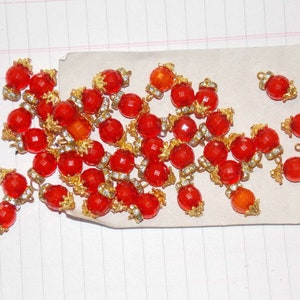 May include: A collection of red glass beads with gold accents and rhinestones. The beads are arranged in a loose pile on a white surface.