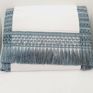 May include: A roll of blue decorative fringe. The fringe features a woven top section and long tassels in a muted blue hue, with a gradient effect. The fringe is wrapped around a white cardboard backing, suitable for craft projects.