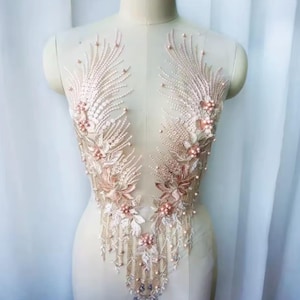 Op de afbeelding: Een delicate, perzikroze kanten applicatie met ingewikkelde bloemen- en kralendetails. Het ontwerp heeft gelaagde bloemen, parelaccenten en bungelende kralenstrengen, waardoor een elegant, sierlijk stuk voor versiering ontstaat.