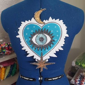 May include: A turquoise blue heart-shaped patch with a black and white eye in the centre. The eye has a blue and white pupil. The patch is surrounded by white sequins and has a gold crescent moon and a gold star at the top and bottom.