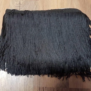 Op de afbeelding: Close-up van een zwarte franjeband. De band bestaat uit talrijke lange, dunne strengen, die een gelaagd textureffect creëren. De franje is effen zwart, met een geweven band aan de bovenkant. Geschikt voor kleding of woondecoratie.