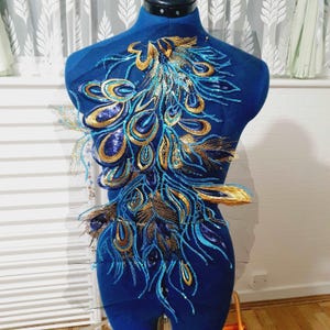Könnte beinhalten: Blaue und goldene Pfauenfeder-Applikation mit Pailletten. Diese dekorative Verzierung ist perfekt, um Kleidung, Accessoires oder Heimtextilien einen Hauch von Eleganz zu verleihen.
