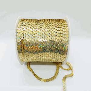 Puede incluir: Un carrete de ribete de lentejuelas doradas. El ribete está hecho de pequeñas lentejuelas redondas que están cosidas juntas en una línea continua. Las lentejuelas tienen un ligero brillo iridiscente.