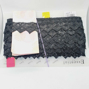 Op de afbeelding: Zwarte kantbies met een geometrisch patroon. De bies is verpakt met een wit label waarop "Essential", "Trims", "Beads", "Cords", "Laces" staat en een roze label waarop "Essential Trims" staat.