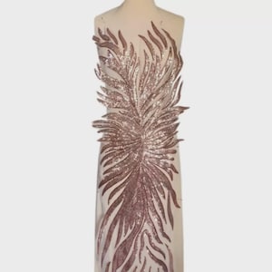 Może przedstawiać: Przezroczysta tkanina z dużym, cekinowym wzorem pióra w odcieniach różowego złota i srebra. Szczegółowy wzór jest umieszczony centralnie, tworząc uderzający efekt wizualny. Cekiny odbijają światło, dodając tkaninie odrobinę blasku.