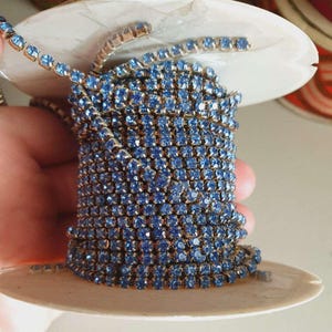 Può includere: Un rotolo di passamaneria a catena di strass blu chiaro. La catena è composta da piccoli strass quadrati collegati da anelli metallici.