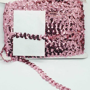 May include: A roll of pink sequin trim. The trim is made of small, round sequins that are sewn together in a continuous line. The sequins are a light pink color and have a slight shimmer to them.