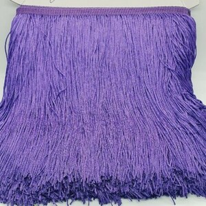 May include: A vibrant purple fringe trim featuring a dense row of long, thin, hanging threads attached to a woven header tape. The fringe is neatly wound around a white cardboard spool, displaying a consistent, uniform colour and texture throughout.