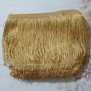 May include: Gold fringe trim with a woven texture. The fringe is approximately 2.5 cm long.