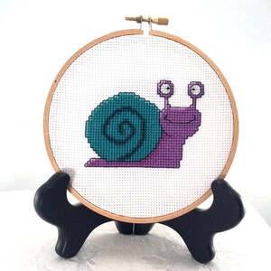 May include: A purple and teal snail cross-stitch embroidery in a wooden embroidery hoop on a black stand.