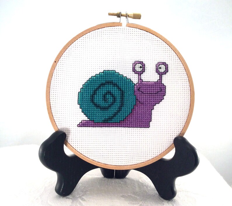 May include: A purple and teal snail cross-stitch embroidery in a wooden embroidery hoop on a black stand.