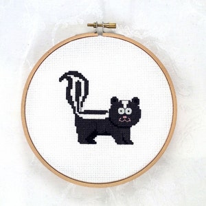 May include: A cross-stitch embroidery of a skunk in a wooden hoop. The skunk is black and white with a fluffy tail and a pink mouth. The background is white.