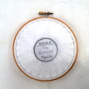 May include: A close-up of the back of a round wooden embroidery hoop with white fabric stretched across it. The fabric has a hand-stitched border and a black and white label that reads "Handmade by The Compass Needle".