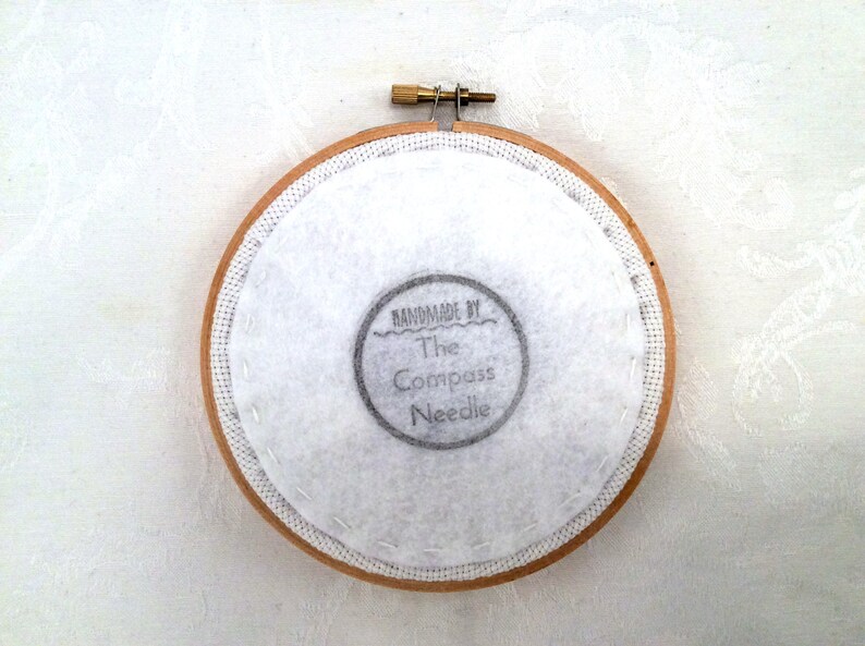 May include: A close-up of the back of a round wooden embroidery hoop with white fabric stretched across it. The fabric has a hand-stitched border and a black and white label that reads "Handmade by The Compass Needle".