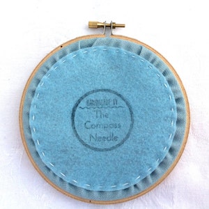 May include: The back of a round embroidery hoop with a light blue fabric backing. The fabric has a stamp that reads "Handmade by The Compass Needle".