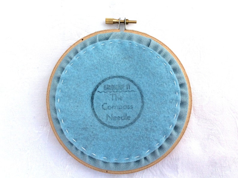 May include: The back of a round embroidery hoop with a light blue fabric backing. The fabric has a stamp that reads "Handmade by The Compass Needle".