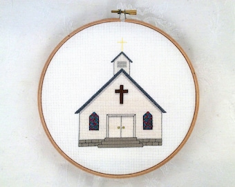 May include: Cross-stitch embroidery of a white church with a brown cross and steeple. The church has a gray roof and stained glass windows. The embroidery is set in a wooden hoop.