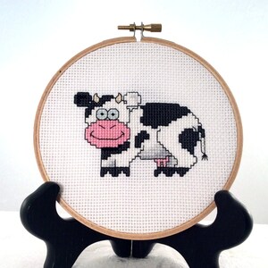 May include: A white and black cow with pink cheeks and big eyes is embroidered on a white canvas in a round wooden embroidery hoop.