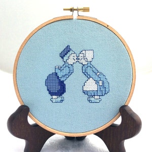 May include: A blue and white cross-stitch embroidery of two figures kissing, framed in a wooden hoop with a brown stand.