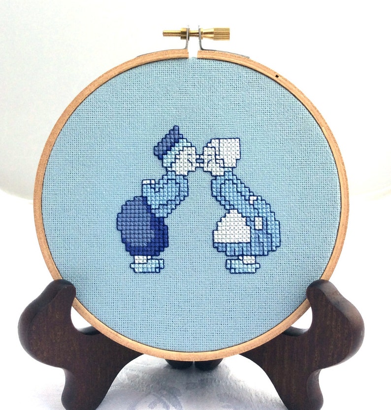 May include: A blue and white cross-stitch embroidery of two figures kissing, framed in a wooden hoop with a brown stand.