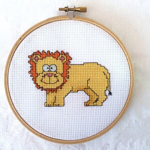 Puede incluir: Un aro de bordado de punto de cruz con un león amarillo con una melena marrón y orejas rosas. El león tiene una cara sonriente con ojos negros y una nariz rosa.