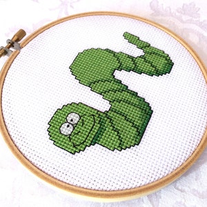 May include: A green worm with a smiling face is embroidered on a white fabric stretched over a wooden embroidery hoop.
