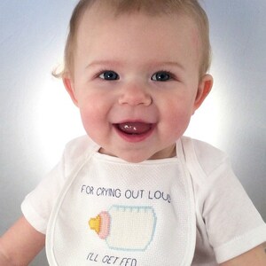 Op de afbeelding: Een baby draagt een witte slabbetje met een borduursel van een babyflesje en de tekst "For crying out loud I'll get fed".