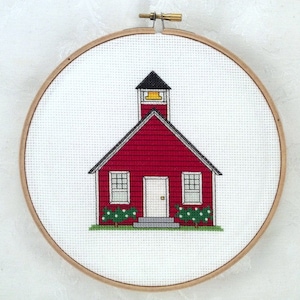 May include: A red schoolhouse with a yellow bell tower, white windows and door, and green bushes on either side, embroidered on white fabric in a wooden embroidery hoop.