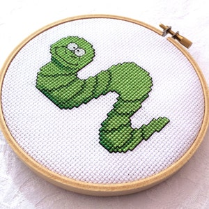 May include: A green embroidered worm with a smiling face is stitched on a white fabric stretched over a wooden embroidery hoop.
