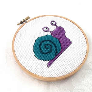 May include: A purple and teal snail cross-stitch pattern in a wooden embroidery hoop.