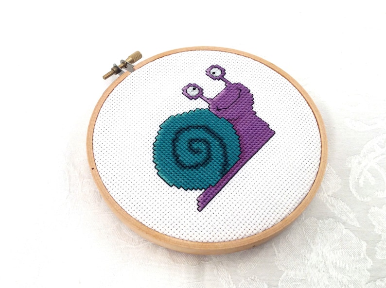 May include: A purple and teal snail cross-stitch pattern in a wooden embroidery hoop.