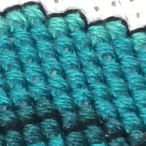May include: Close-up of a teal blue cross-stitch fabric with a black outline. The fabric is textured and has a woven appearance.