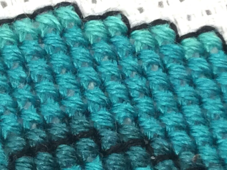 May include: Close-up of a teal blue cross-stitch fabric with a black outline. The fabric is textured and has a woven appearance.