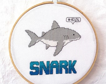 May include: A white embroidery hoop with a gray and white pixelated shark with a speech bubble that says "#@$%". The word "SNARK" is embroidered in blue below the shark.