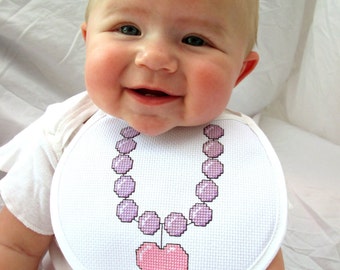 May include: A baby wearing a white bib with a pink heart and purple bead necklace embroidered on it.