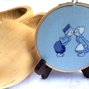 May include: A blue and white cross-stitch embroidery of two people kissing, framed in a wooden hoop, with a pair of wooden clogs in the background.