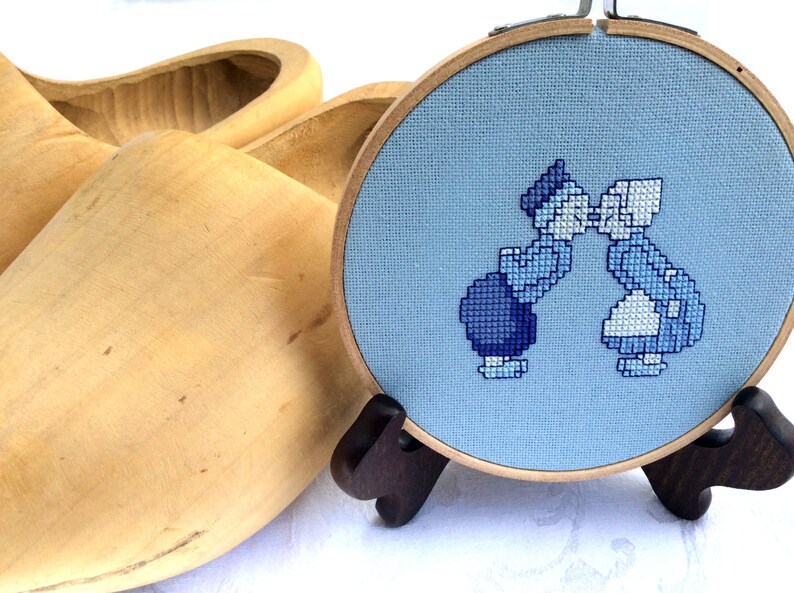 May include: A blue and white cross-stitch embroidery of two people kissing, framed in a wooden hoop, with a pair of wooden clogs in the background.