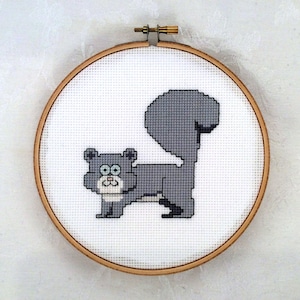 May include: A cross-stitch embroidery of a grey cat with a large, fluffy tail, set in a wooden hoop. The cat is depicted in a pixelated style, with a white chest and large, round eyes. The background is white.