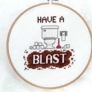 May include: White embroidery hoop with a brown and white cross-stitch design. The design features a toilet, toilet paper, a plunger, and the text "Have a Blast".