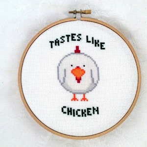 May include: A white embroidery hoop with a cross-stitch design of a white chicken with a red comb and orange beak. The text "Tastes Like Chicken" is stitched in black above and below the chicken.