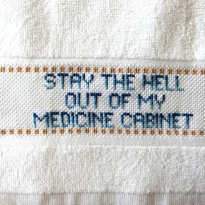 Op de afbeelding: Witte handdoek met blauwe geborduurde tekst die "Stay the hell out of my medicine cabinet" luidt.