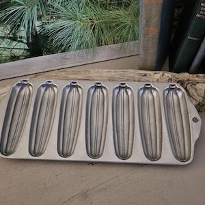 May include: A vintage, rectangular, cast-iron cornbread mold with seven individual, elongated, ribbed cavities. The mold is made of silver-colored metal and sits on a neutral-toned surface. The background includes a window and wooden crates.
