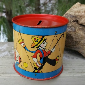 May include: A vintage tin piggy bank with a blue and yellow design featuring a cartoon character wearing a sombrero and holding a stick. The piggy bank has a red lid with a slot for coins.