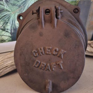 May include: A circular, dark brown, weathered cast iron check draft door featuring a hinged top mechanism and mounting holes on the sides. The words CHECK DRAFT are embossed in raised capital letters across the center of the metal surface.