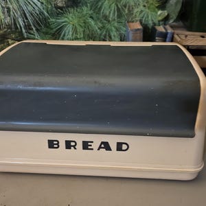 May include: A vintage bread box with a cream-colored base and a dark gray lid. The word "BREAD" is printed in bold black letters on the front. The box has a rounded top and a rectangular shape.